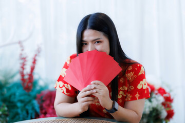Portrait of happy asian woman relaxing wearing red cheongsam dress holding red envelopes at home in the living room indoor decoration for Chinese new year festival celebrate culture of china holiday
