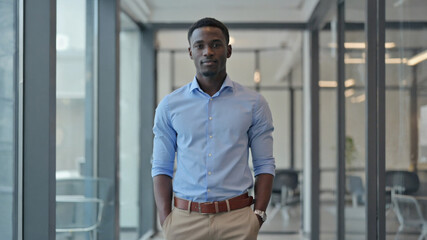 A young man standing in a modern office, wearing a sweater and trousers against a backdrop of bookshelves and decor.