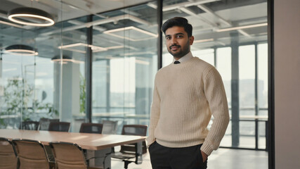 A young man standing in a modern office, wearing a sweater and trousers against a backdrop of bookshelves and decor.