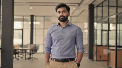 A young man standing in a modern office, wearing a sweater and trousers against a backdrop of bookshelves and decor.