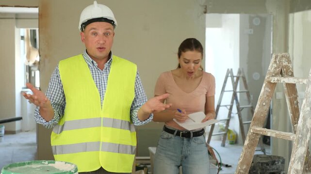 Portrait of frustrated workman wearing protective uniform and holding papers standing at renovating object, having problems with renovation, woman house painter standing on background. High quality 4k