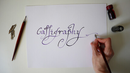 Top view of modern script calligraphy creation process. The word Calligraphy written with a pen, ink bottles, and nibs on a clean white background. Womens hand. Ideal for art and creative themes.