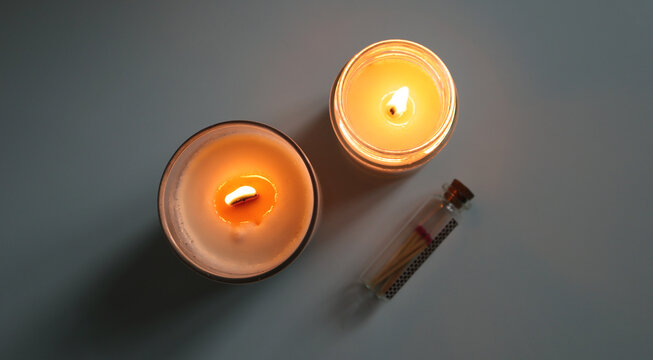 Top view of two burning scented candles in glass jars and a small glass bottle with decorative matches. The soft, warm light creates an atmosphere of coziness, spa relaxation, and home meditation. - Powered by Adobe
