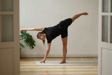 A young woman in black athletic wear performs the half moon pose (Ardha Chandrasana) on a grey mat, balancing on one leg and hand in a minimalist room with wooden parquet floors