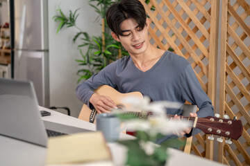 Asian handsome man strumming acoustic guitar in living room at home. 