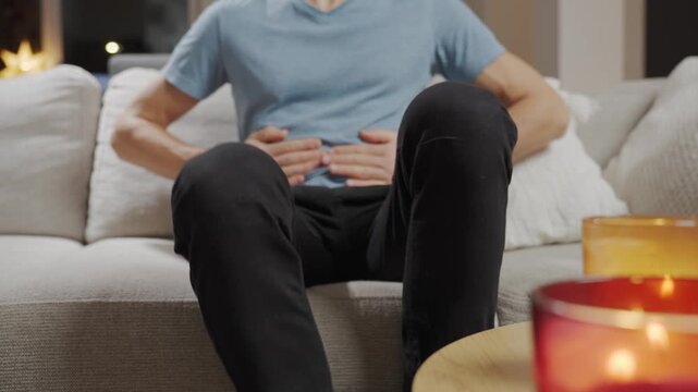 Man sitting on sofa in living room holding stomach and suffering from abdominal pain or cramps. Person suffering from health issues, digestion problems or food poisoning at home