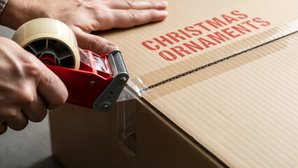 Close-up of hand using tape dispenser on brown cardboard Christmas ornaments box. Moving box contains Christmas ornaments for storage, ready for holiday season.