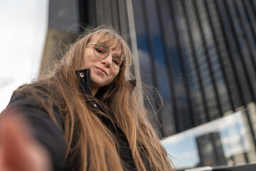 Fototapeta premium Woman with long hair and bangs wearing glasses, capturing a low-angle self-portrait outdoors in a city setting
