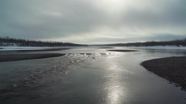 Wide winter river landscape with sun reflection on water dark gravel bars snow-covered banks and bleak overcast sky.