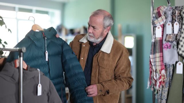 Senior man chooses a warm down jacket among the assortment of outerwear. Pensioner stands near a coat rack with jackets and looks at his size. High quality 4k footage