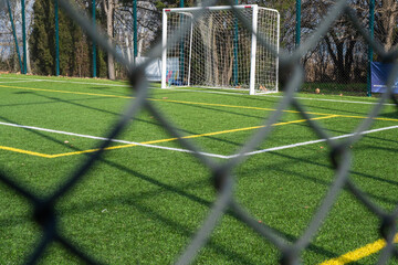 a view through a blurred gray chain-link fence of a white soccer goal standing on a green...