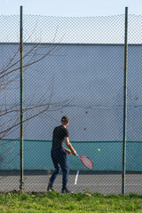 Fototapeta premium a male tennis player hitting a yellow ball against a practice wall on a hard court, viewed through a chain-link fence.