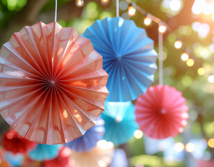 Vibrant paper fans in orange, blue, and red hang outdoors, illuminated by soft sunlight and bokeh string lights, creating a cheerful, festive atmosphere.