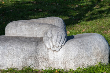 a detail of a rough gray stone sculpture resting on a grassy lawn, depicting a stylized arm and hand clasping together in bright sunlight.