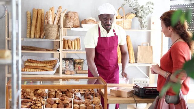 Man baker packaging croissants in a plastic bag, serving a woman customer in a bakery filled with fresh pastries.