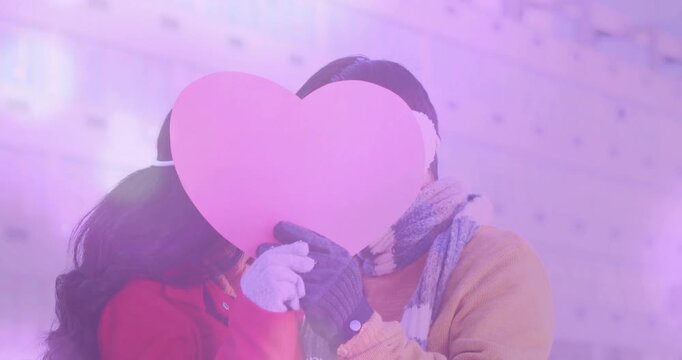 Couple stepping onto balcony, holding pink heart prop and kissing behind it for marketing