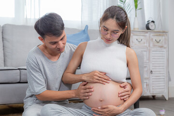 Asian pregnant woman and man sitting on floor, both cradling belly and smiling lovingly. love and anticipation of their baby. Cozy home setting. family or parenthood togetherness activities concept