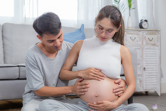 Asian pregnant woman and man sitting on floor, both cradling belly and smiling lovingly. love and anticipation of their baby. Cozy home setting. family or parenthood togetherness activities concept - Powered by Adobe