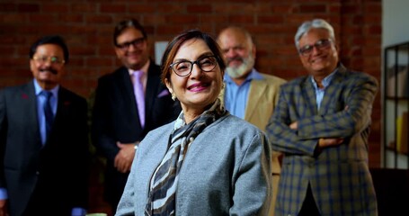Portrait of Indian senior businesswoman leading team as senior Indian Asian female executive smiles at camera with confident expression while colleague stands behind her in modern corporate office