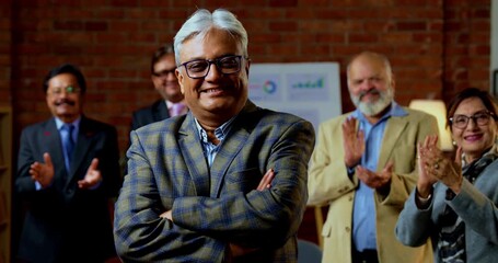 Portrait of Indian senior businessman leading team as senior Indian Asian male executive smiles at camera with confident expression while colleague stands behind him in modern corporate office - Powered by Adobe
