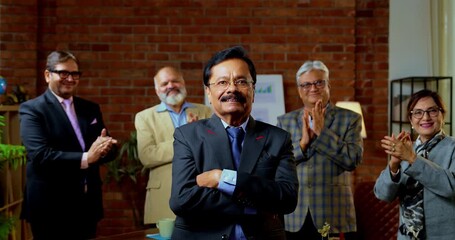 Portrait of Indian senior businessman leading team as senior Indian Asian male executive smiles at camera with confident expression while colleague stands behind him in modern corporate office