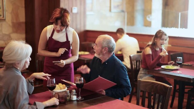 Positive female server listening carefully while standing beside old couple enjoying evening meal in restaurant