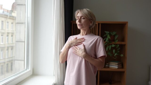 Positive middle aged woman do breathing exercise for relaxation and mental health during day at home, inhaling exhaling, relieving stress. Female stands beside window and breathing with closed eyes.