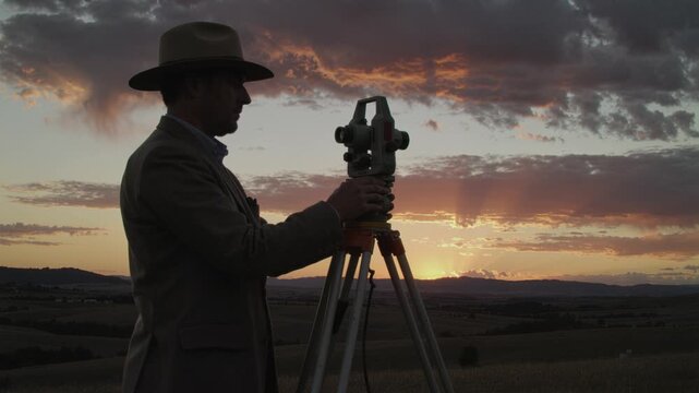 Silhouette of a land surveyor using a theodolite on a tripod against a dramatic sunset sky over rural hills, suitable for engineering and construction concepts.