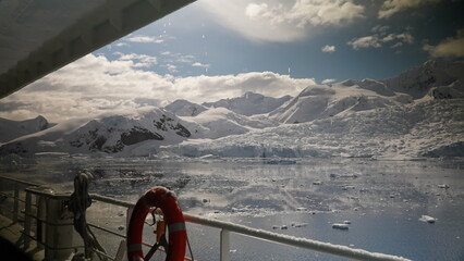Antarctic view from ship ,iced  railing and life buoy in foreground with ice drifting in the ocean and snow mountains in the background reflecting in the ocean with blue skies and thick white clouds © Amelia