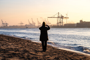A person standing at the beach at the Elbe River in Hamburg taking photo of the sunrise over the harbor