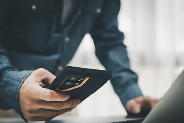 Man holding a smartphone while working on a laptop. Concept of digital multitasking, mobile banking, search seo marketing trends and wireless communication in a professional or home office environment