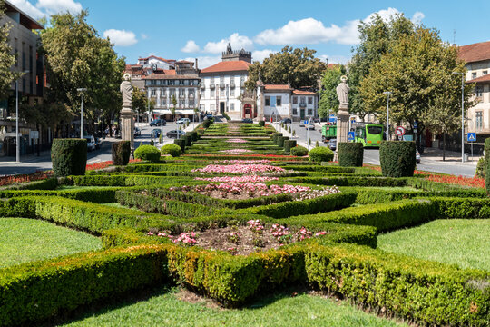 Jardim em Guimar&atilde;es com arbustos verdes em padr&otilde;es geom&eacute;tricos, canteiros floridos e a distinta arquitetura portuguesa de edif&iacute;cios hist&oacute;ricos e est&aacute;tuas sob um c&eacute;u azul de ver&atilde;o