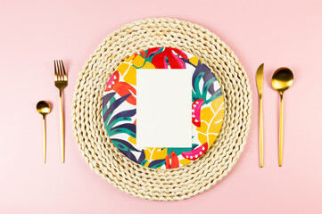 Top view of brown wicker table mat, colorful plate, gold cutlery on pink background. White card,...