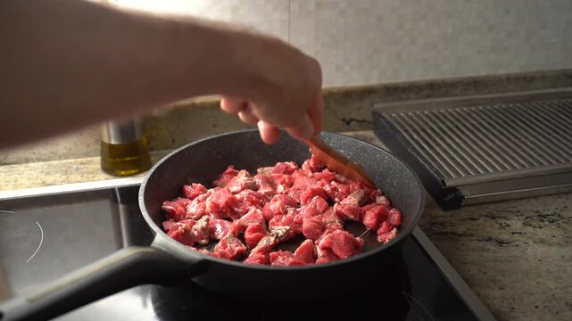 Close-up of raw beef cubes being fried in a pan with a wooden spatula, cooking process in a home kitchen.