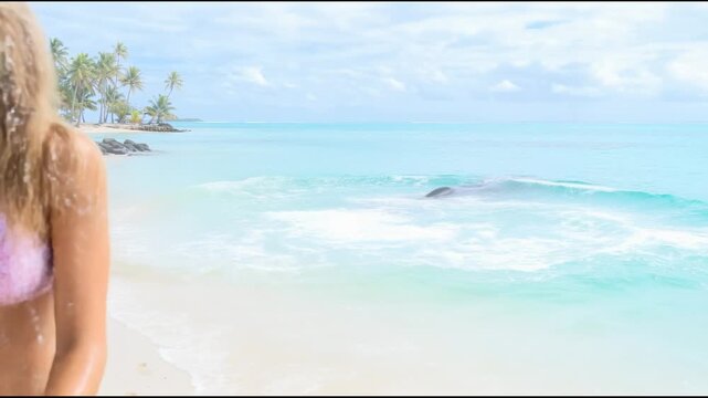 Girl in bikini walks along beach waving hand while whale in background slaps tail on water raising fountain of spray
