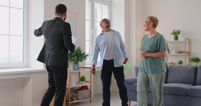 Realtor leads elderly clients on an apartment tour, discussing contract terms, purchase details in a bright living room. Real estate agent guides senior couple through apartment viewing.