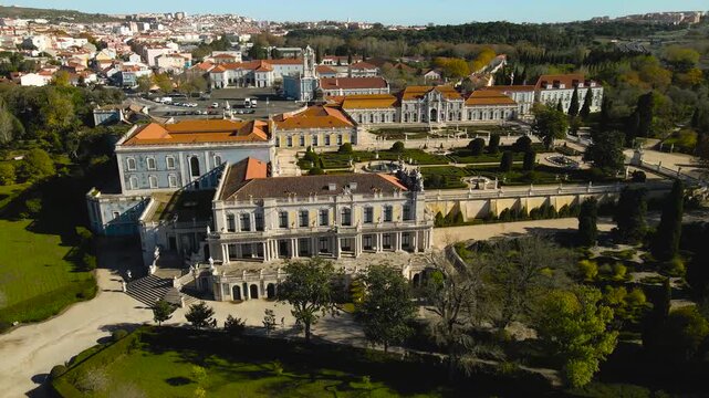 Aerial backward reveal of Queluz National Palace in Portugal showing formal French gardens, fountains, trimmed hedges and baroque architecture on a sunny day, filmed with a drone, no people visible.