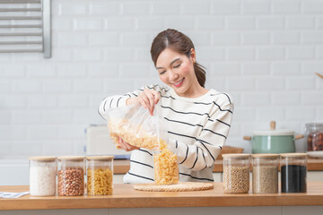 Young asian woman smiling while pouring cornflakes into a glass jar in a bright kitchen, focusing...