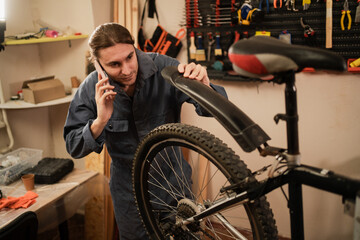 Bike service, repair and upgrade concept. Young man mechanic in a bike workshop holding smartphone and discussing while working on bicycle