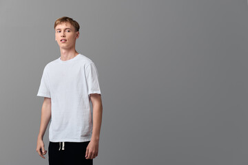Young man in a white t shirt stands in a studio, casual wear and relaxed pose against a gray...