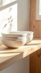 Stacked White Ceramic Bowls Catching Warm Sunlight on a Wooden Shelf Near a Window