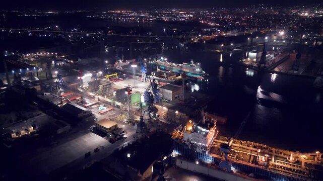 Night aerial video of a ship repair yard with dry docks, cranes and vessels illuminated by industrial lights. Large shipyard operating after dark with maritime infrastructure and harbor waters