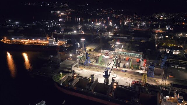 Night aerial video of a ship repair yard with dry docks, cranes and vessels illuminated by industrial lights. Large shipyard operating after dark with maritime infrastructure and harbor waters
