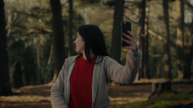  a young woman hiking in a forest as she video calls with her smartphone. With a bright smile and a backpack, she captures the moment amidst the natural scenery, reflecting the joy 