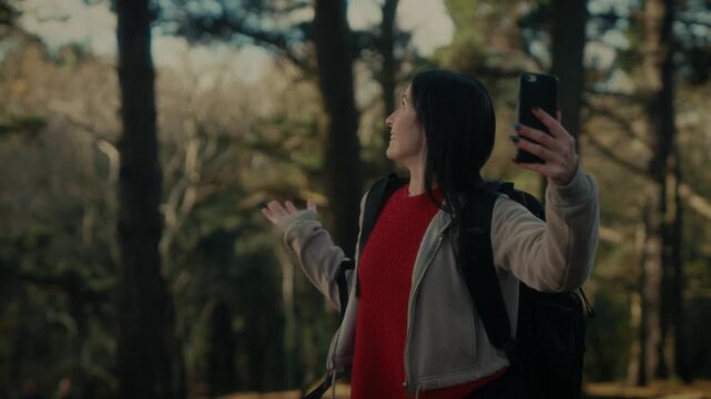  a young woman hiking in a forest as she video calls with her smartphone. With a bright smile and a backpack, she captures the moment amidst the natural scenery, reflecting the joy 
