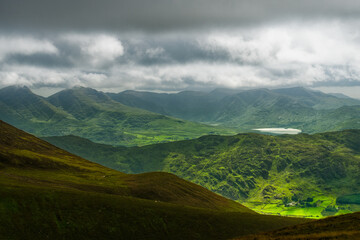 A wide, daytime scene features a green mountain landscape in Killarney National Park, Ireland, with moody skies above the rolling hills. A lake is seen in the distance.