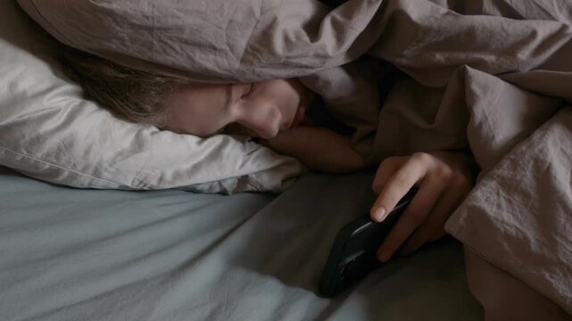 Depressed Teenage Boy Hiding Under Blanket with Smartphone in Dark Bedroom. Conceptual Visual of Youth Mental Health Crisis, Loneliness, Social Isolation, and Teen Depression in Digital Age