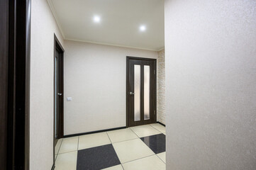 Fototapeta premium Narrow hallway with light beige textured walls, dark wooden doors with frosted glass panels, two-tone tiled floor (beige & dark), recessed ceiling lights, minimalist design