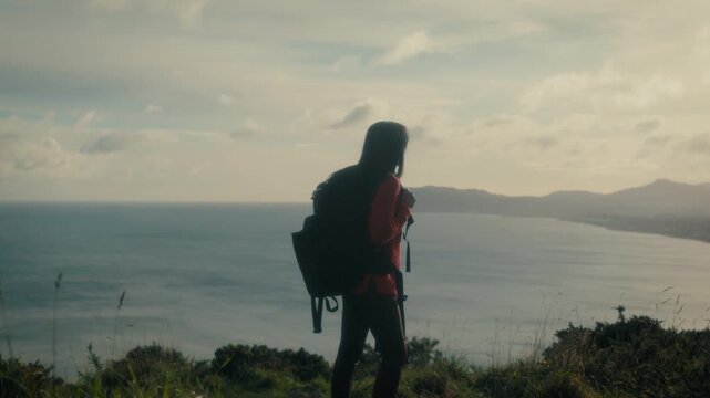  a young woman standing at a scenic viewpoint, gazing out over a tranquil body of water and distant hills. With her backpack slung over her shoulders.