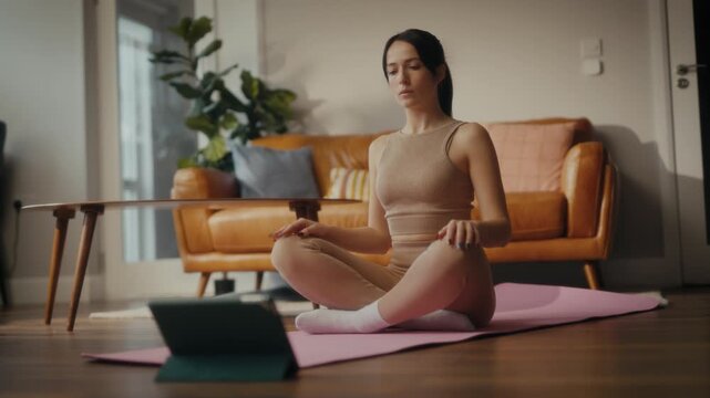 woman practising yoga in a cosy living room setting. She is in a lunging pose on a yoga mat, with a tablet in front of her displaying a workout or meditation session. 